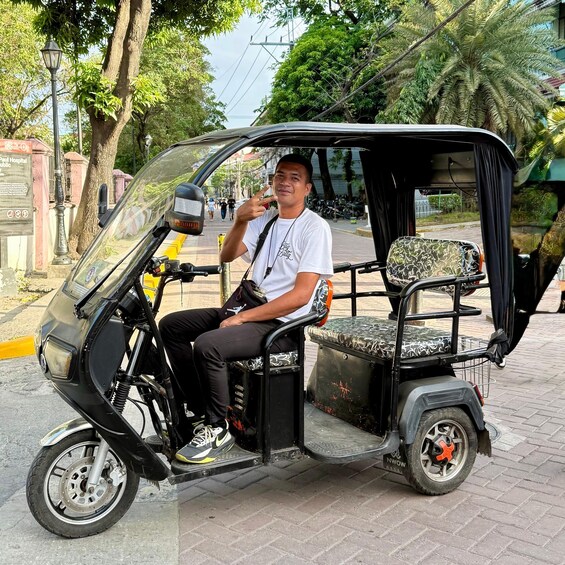 A man sitting in a three-wheeled vehicle with a canopy, parked on a paved street with trees and a sign in the background. 1