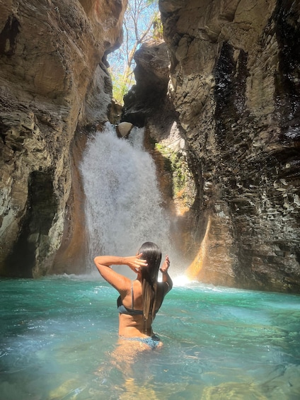 A person standing in clear water with a waterfall in a rocky cave. 4