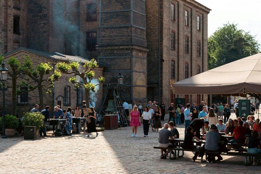 En brostensbelagt plads med mennesker, der spiser ved borde, en murstensbygning og et overdækket område med et telt. 5