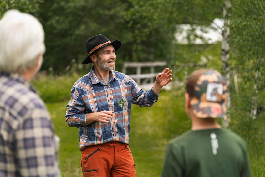 A person in a plaid shirt and hat gesturing, standing in a grassy area with trees and a wooden structure in the background. 5