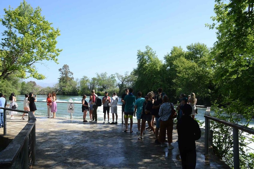 A group of people standing on a concrete walkway by a river, with trees and a clear sky in the background. 5