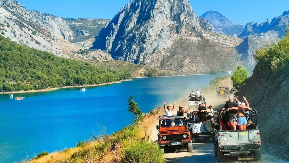 A group of people in jeeps and a boat on a lake surrounded by mountains. 1