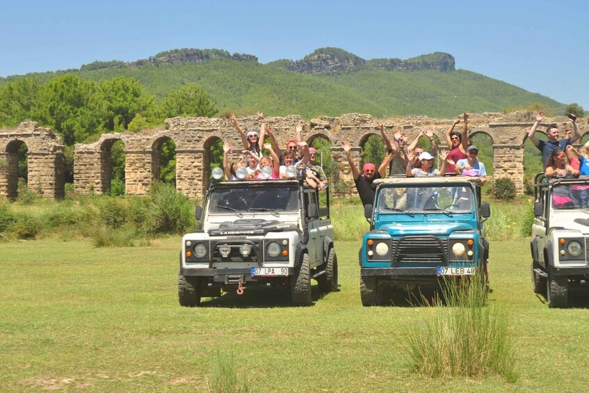 Three off-road vehicles with passengers, driving on a grassy field with historical stone ruins in the background. 2