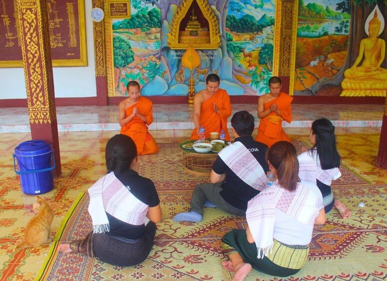Monks in orange robes seated in a circle, with a mural depicting a serene landscape in the background. 1