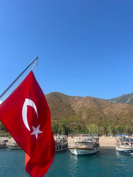 A red flag with a white crescent and star, a body of water, boats, a beach, and mountains in the background. 3