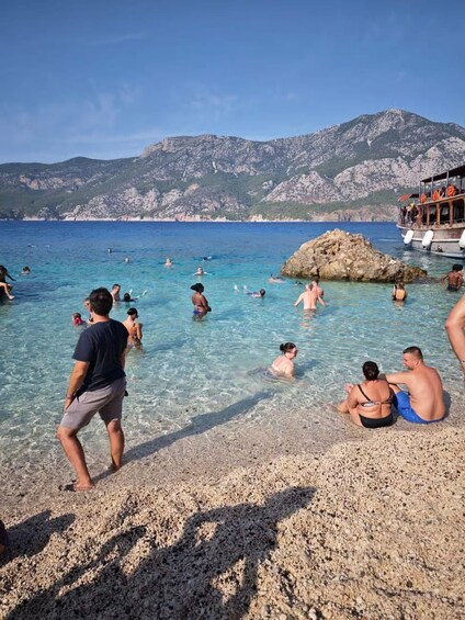 A beach with clear water, people swimming and sunbathing, and rocky mountains in the background. 2