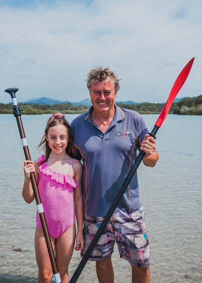 A man and a child holding paddles by a lake with mountains in the background. 2