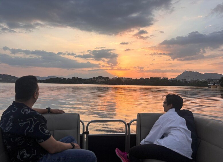 Two people seated on a boat, facing a sunset over a body of water with distant hills. 1