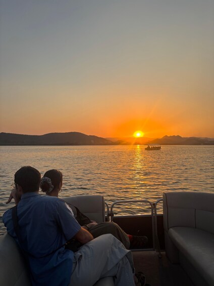 Two people sitting on a boat, watching a sunset over a calm body of water with a distant boat visible. 4