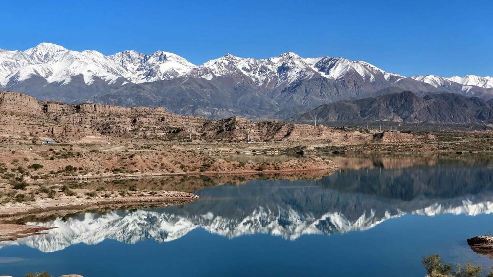 A calm lake reflecting snow-capped mountains, rocky terrain, and sparse vegetation. 1