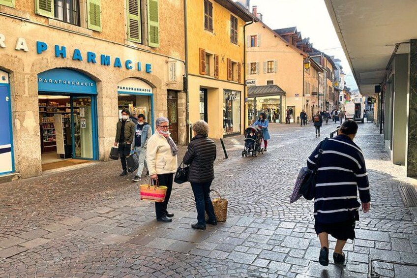 A cobblestone street with people walking, a pharmacy, and buildings with signage. 3