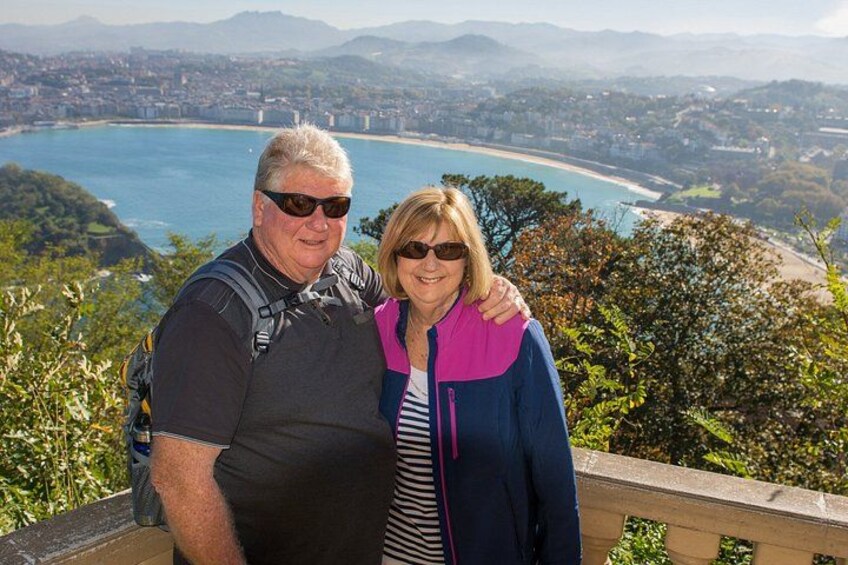 A couple standing on a viewpoint overlooking a coastal city with a beach and mountains in the background. 1