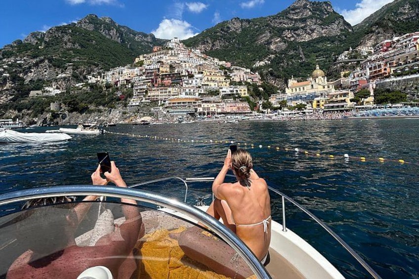 A boat with two people, one taking a photo, and a coastal town with buildings and mountains in the background. 5