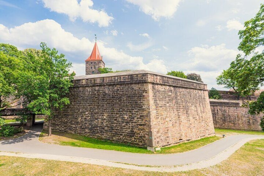 Eine steinerne Festung mit einem Turm mit rotem Dach, umgeben von Grünflächen und einem gepflasterten Weg. 5