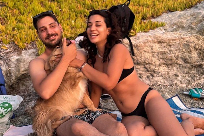 Two people sitting on a beach with a dog, wearing swimwear, and a backpack. 3