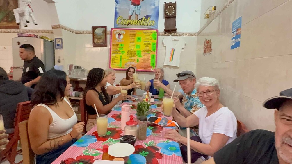 A group of people seated at a table with a colorful floral tablecloth, engaged in conversation and holding drinks. 1