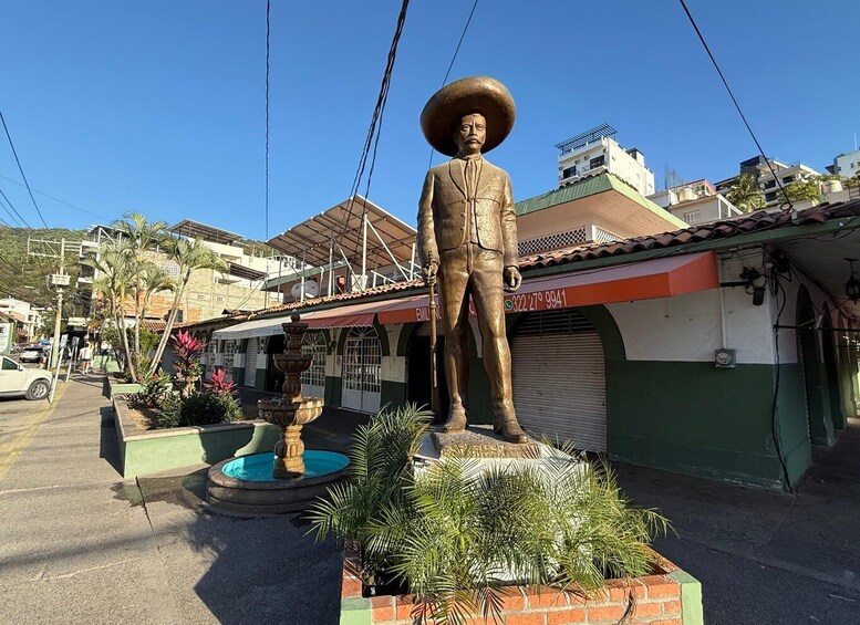 A statue of a man with a large hat, a fountain, and buildings in the background. 2