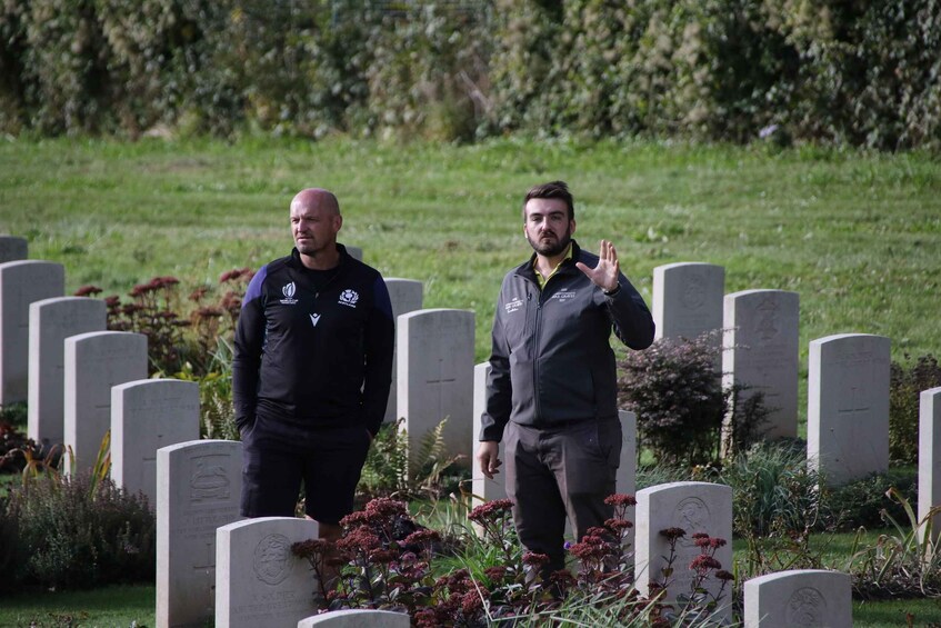 Two individuals walking in a cemetery with headstones and greenery. 4