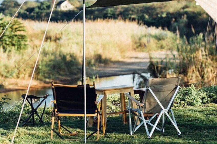 A camping setup with a table and chairs under a tent, a chair on the ground, and a river in the background. 3