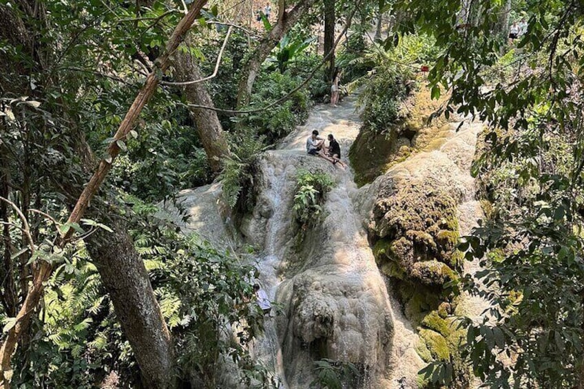 A waterfall in a forested area with people sitting on a rock ledge. 1