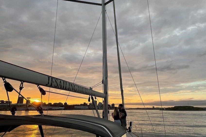 Un velero con 'JFSAILINGTOURISM.COM' en su mástil, una persona de pie en la cubierta y una puesta de sol sobre el agua. 3