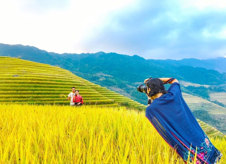 A person taking a photograph of two people in a rice field with mountains in the background. 2