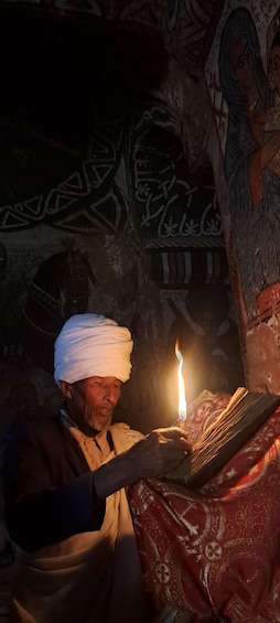 A person with a white head covering, holding a lit candle, in a dimly lit room with patterned walls. 5