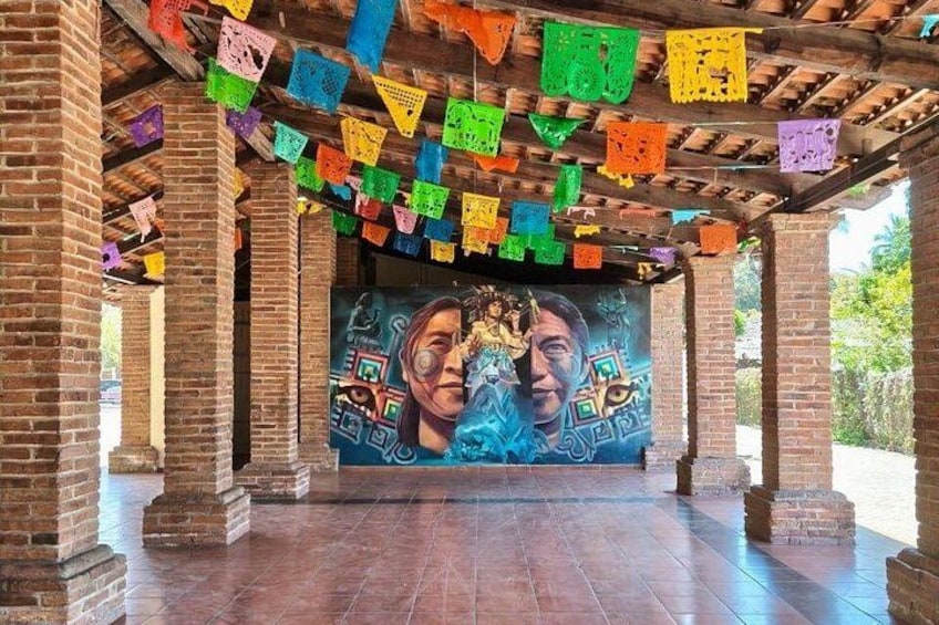 A covered area with colorful paper flags, a mural with geometric patterns, and brick columns. 1