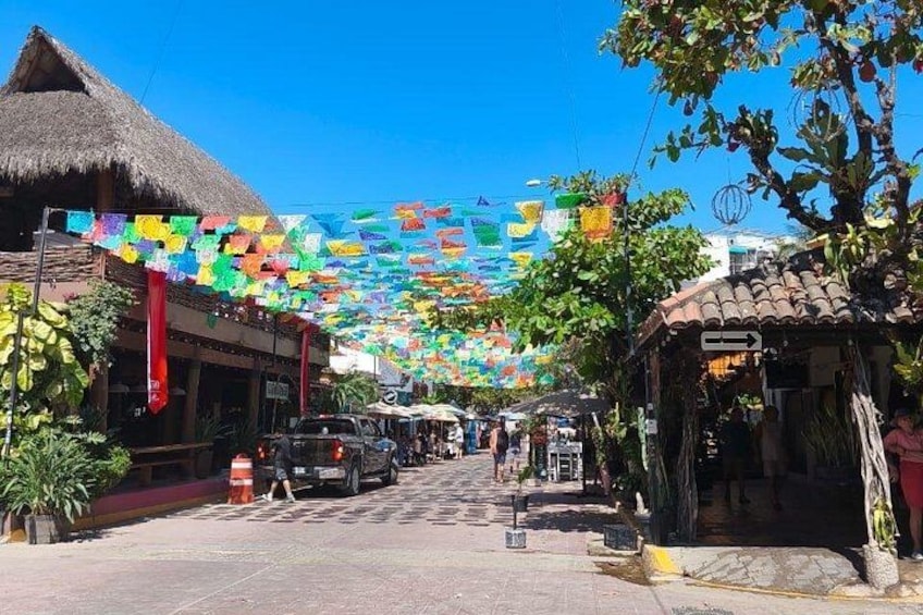 A street lined with buildings, colorful flags, and a thatched-roof structure. 5
