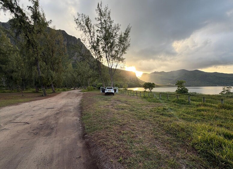 A dirt road with a white car parked on the side, a fenced area, and a lake with mountains in the background. 3