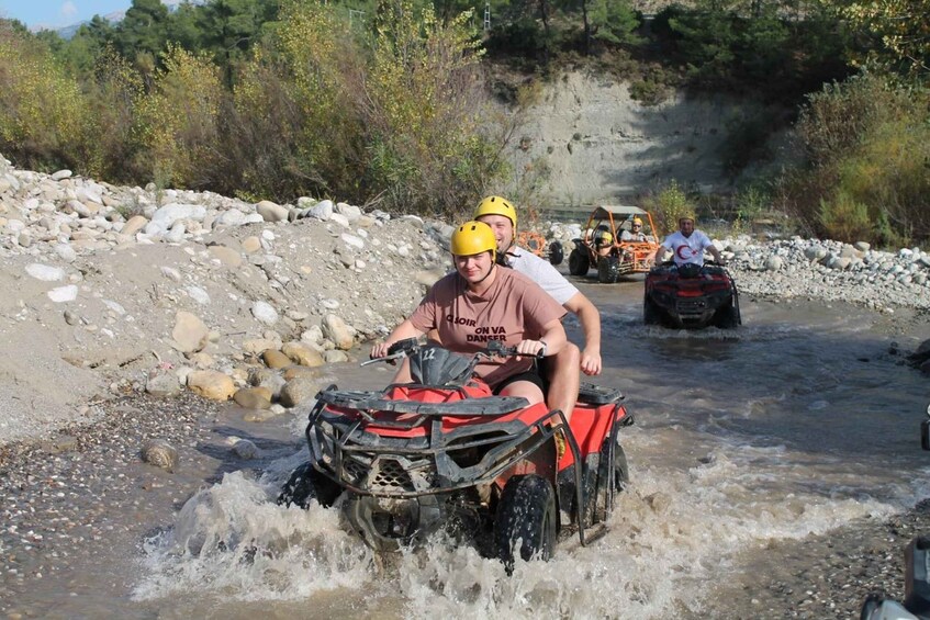 A person riding an ATV through a shallow river surrounded by rocks and vegetation. 1