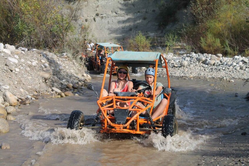 A vehicle navigating a shallow stream with rocky banks and sparse vegetation. 2