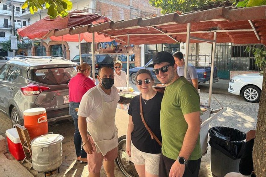 A street scene with people standing under a canopy, a parked car, and a trash bin. 4