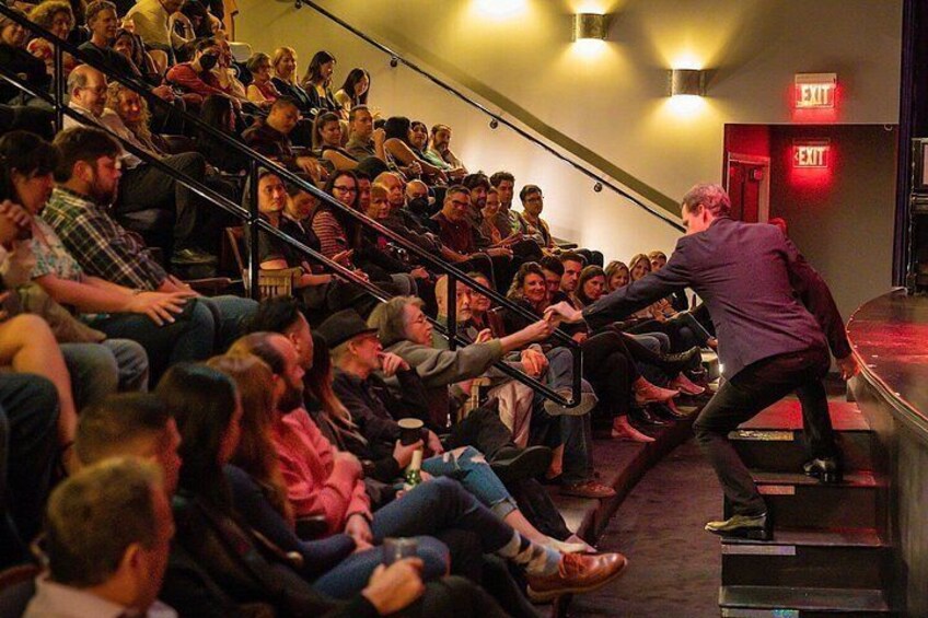Eine Person mit lila Jacke spricht vor einem Publikum, das auf Stufen in einem Theater sitzt. 5