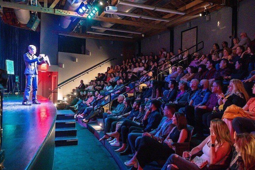Eine Person spricht an einem Rednerpult in einem schwach beleuchteten Auditorium, dessen Zuschauer in Reihen sitzen. 1