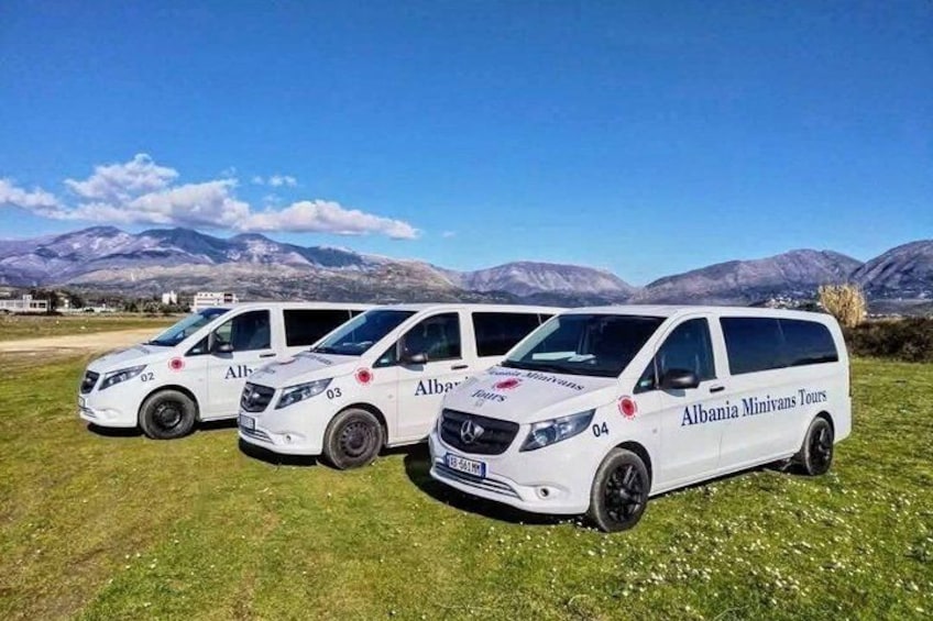Tre vita minibussar med texten ”Albania Minivans Tours” på sidan står parkerade på en gräsmatta med berg i bakgrunden. 1