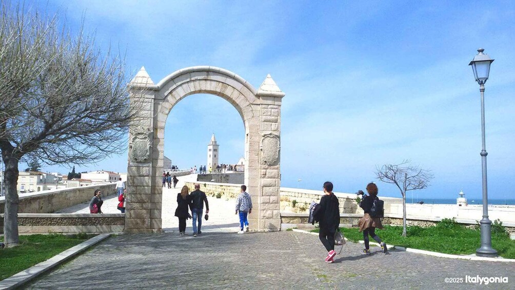 A stone archway with people walking through it, overlooking a coastal area with buildings and a lighthouse. 1