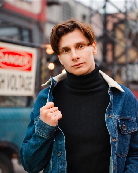 A person wearing a black turtleneck and denim jacket standing in an urban setting with a blurred background featuring a truck and buildings. 3