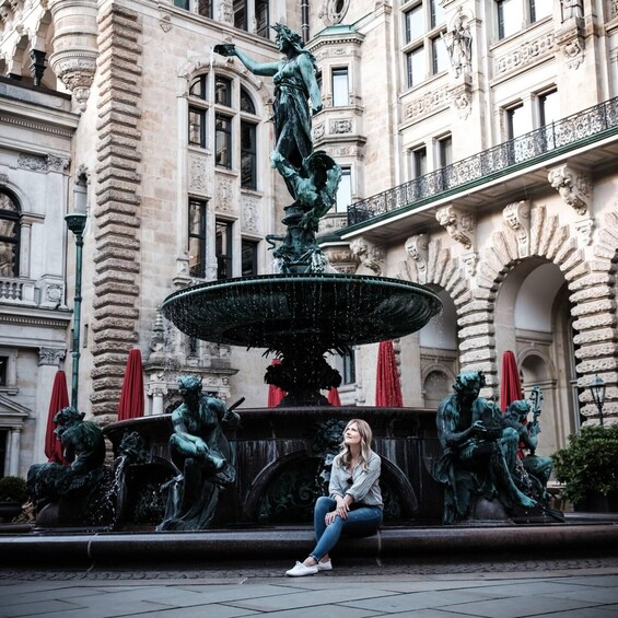 A woman sitting on a bench in front of a historic fountain with statues and a building in the background. 4