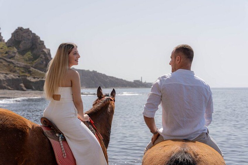 A couple on horseback by the sea with a rocky coastline in the background. 2