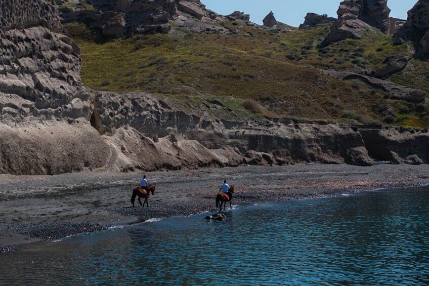 Two people riding horses on a rocky beach with water in the foreground. 1