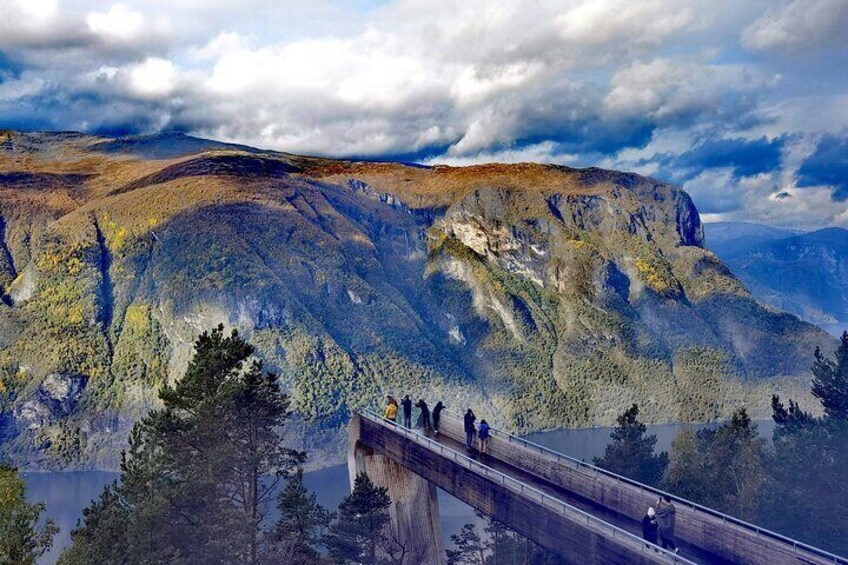 A bridge with people walking, overlooking a mountainous landscape with a lake. 1