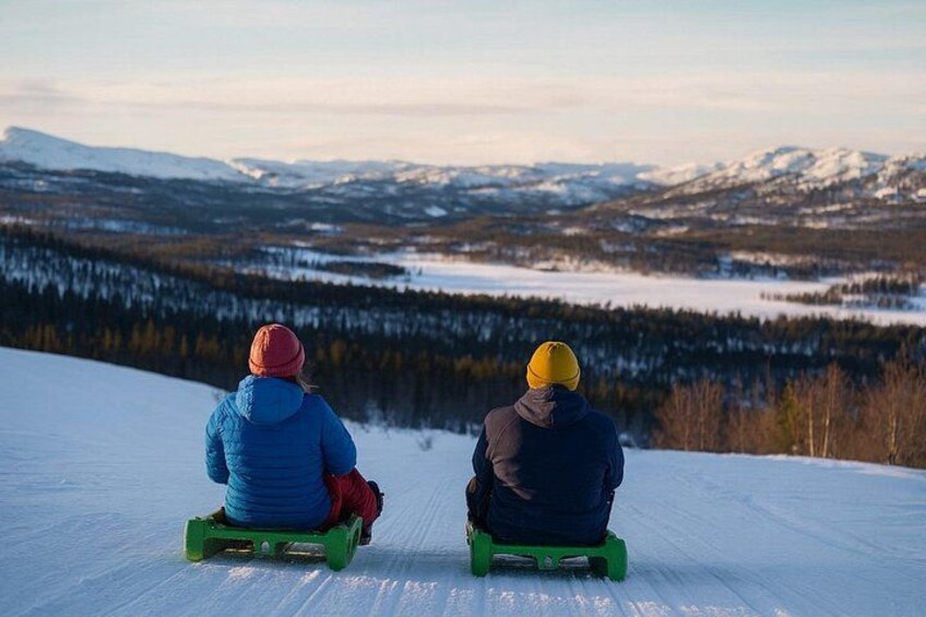 Two people sledding down a snowy slope with a mountainous landscape in the background. 3