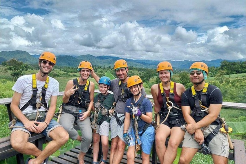 A group of people wearing helmets and harnesses, sitting on a wooden bench with a mountainous landscape in the background. 1