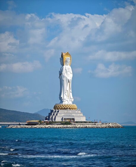 A large white statue with a golden base, standing on a rocky outcrop by the sea. 3