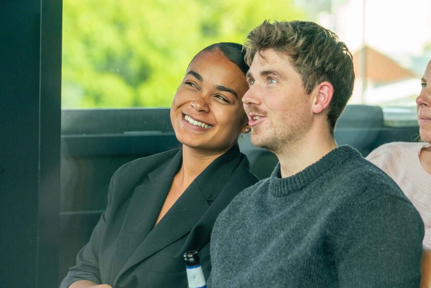 Two people sitting next to each other, one in a black suit and the other in a grey sweater, with a blurred green background. 2
