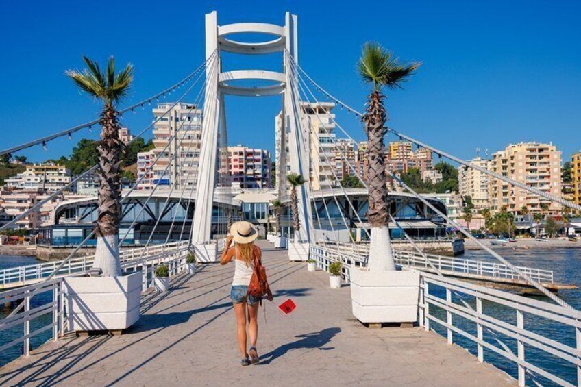 A pedestrian bridge with palm trees, a cityscape in the background, and a clear blue sky. 3