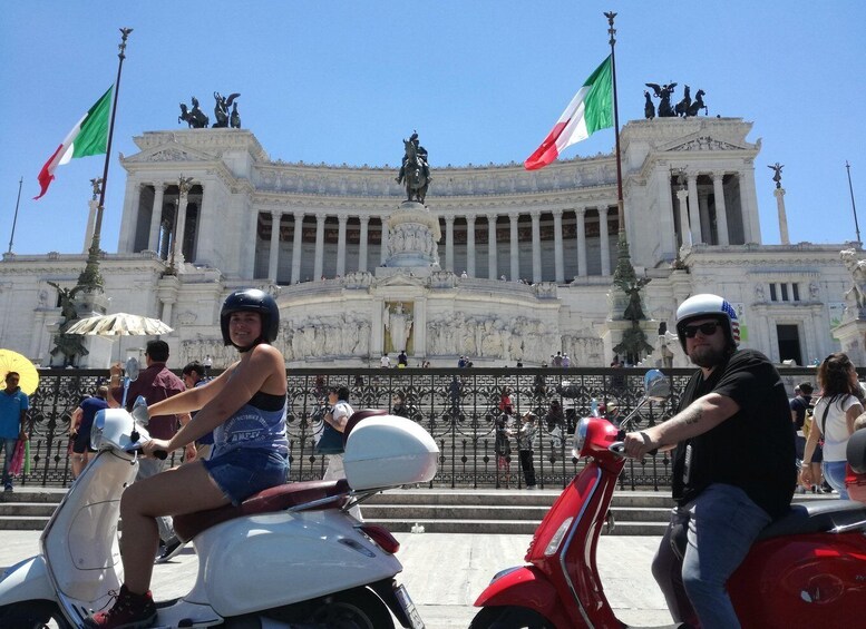 A person on a white scooter and another on a red scooter in front of a grand building with columns and statues, with flags flying. 1