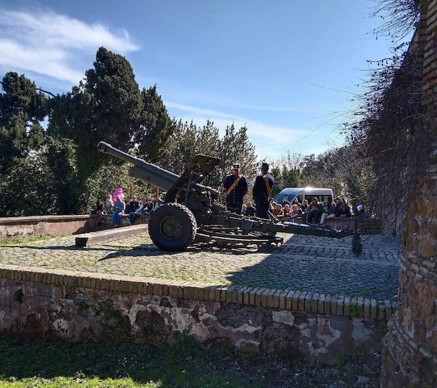 A cannon on display with two uniformed individuals standing beside it, set against a backdrop of trees and a clear sky. 2