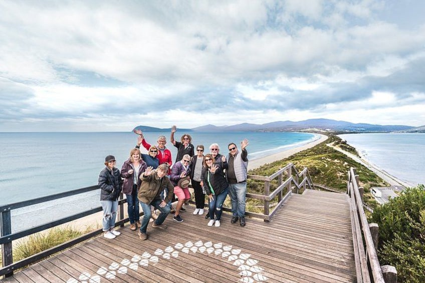 Een groep mensen op een houten looppad met uitzicht op een strand, een heldere zee en verre bergen. 1